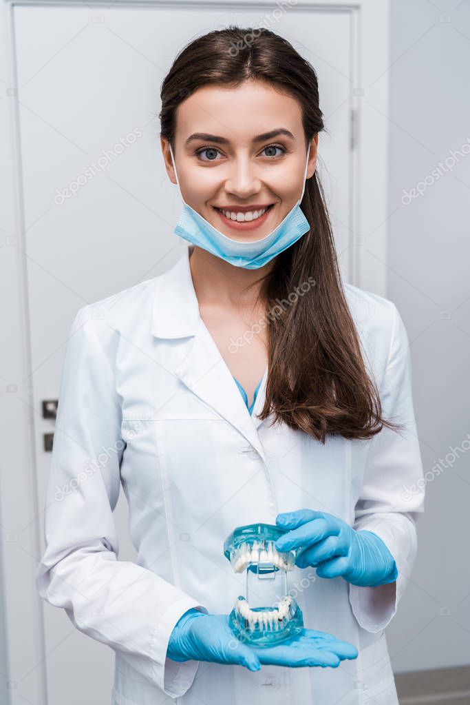 cheerful dentist in medical mask holding teeth model and smiling in clinic 