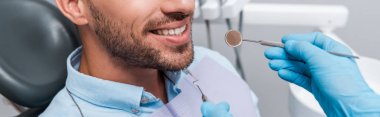 panoramic shot of dentist in latex gloves holding dental instruments near patient  clipart