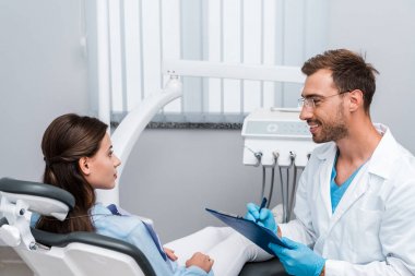handsome dentist in glasses holding pen and clipboard near attractive patient  clipart