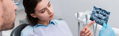 panoramic shot of woman pointing with finger at x-ray near dentist  clipart