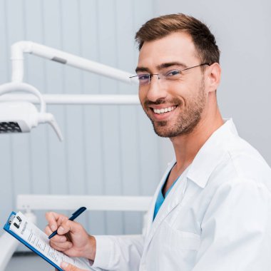 beyaz önlük ve gözlük diş kliniğinde kalem ve pano tutan mutlu sakallı adam 