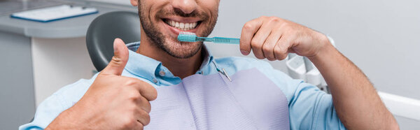panoramic shot of bearded man showing thumb up while holding toothbrush 