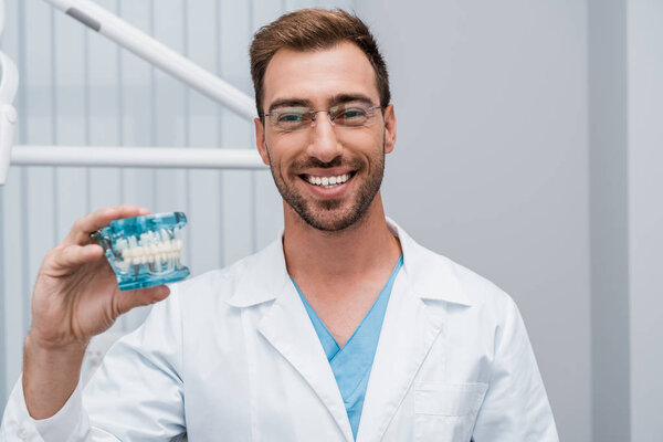 cheerful bearded dentist in glasses holding tooth model in dental clinic 