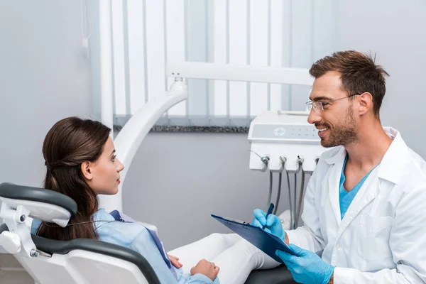 handsome dentist in glasses holding pen and clipboard near attractive patient