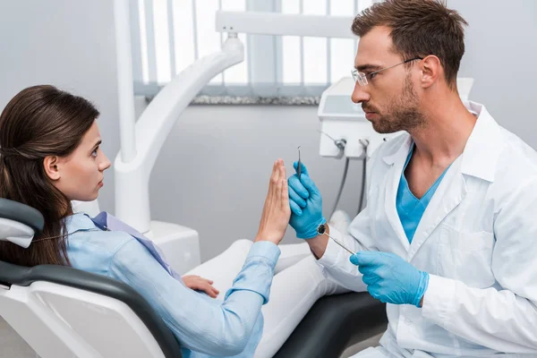 英俊的胡子牙医拿着牙科仪器 看着害怕的女人在诊所打手势 — 图库照片