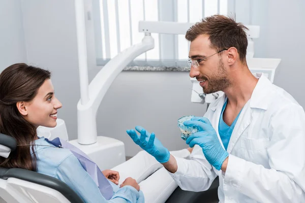 开朗的牙医在眼镜举行牙齿模型 看着幸福的女人 — 图库照片