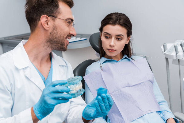 selective focus of handsome bearded man holding retainer and tooth model near attractive patient 