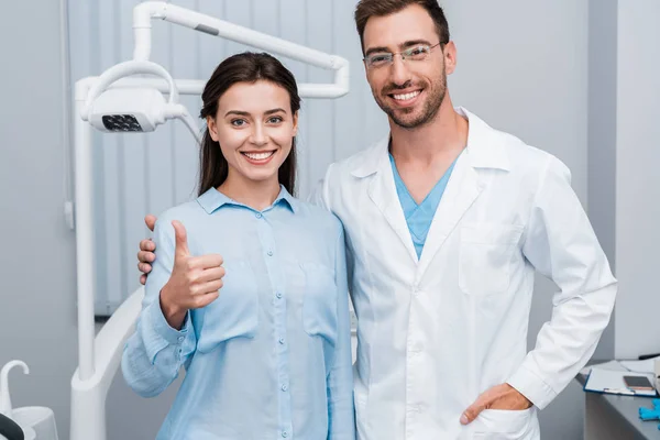 Wesoła Dziewczyna Pokazując Kciuk Pobliżu Przystojny Dentysta Stojący Ręką Kieszeni — Zdjęcie stockowe