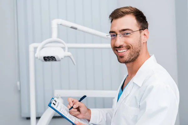 英俊的胡子男子在牙科诊所拿着笔和剪贴板的白色外套和眼镜 — 图库照片