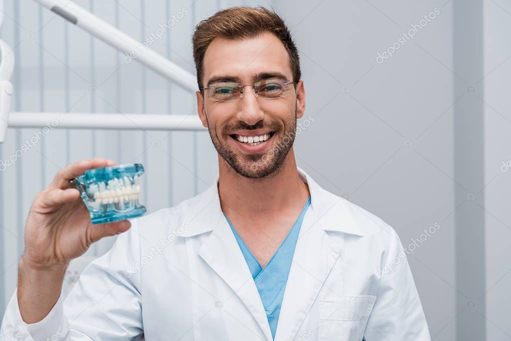 cheerful bearded dentist in glasses holding tooth model in dental clinic 