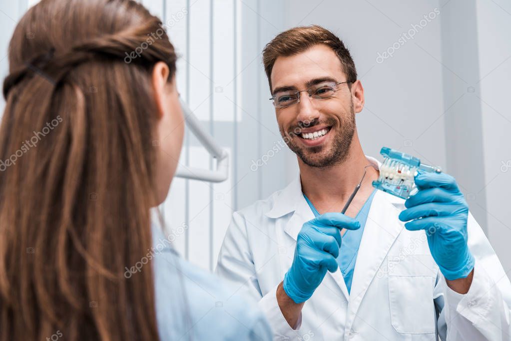 selective focus of happy dentist in glasses holding tooth model near patient