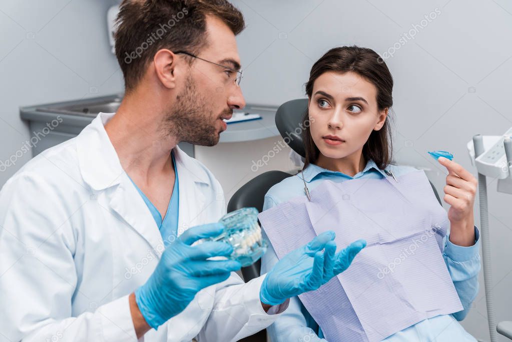 selective focus of handsome dentist in glasses gesturing near woman with retainer 