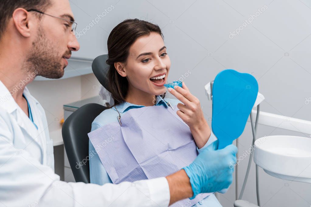 selective focus of dentist in glasses holding mirror near attractive woman with retainer 