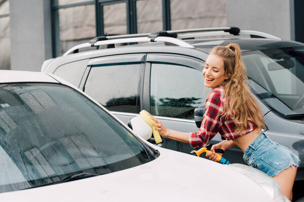 happy woman in denim shorts smiling while washing auto 