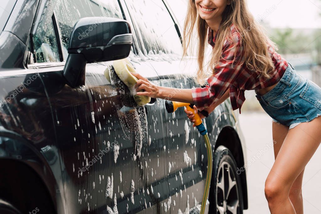 cropped view of positive girl washing wet car in foam 