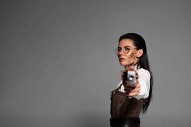 selective focus of confident steampunk woman aiming with pistol at camera isolated on grey clipart