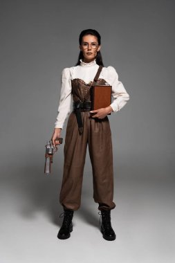 gri üzerinde kitap ve vintage tabanca tutan steampunk kadın tam uzunlukta görünümü