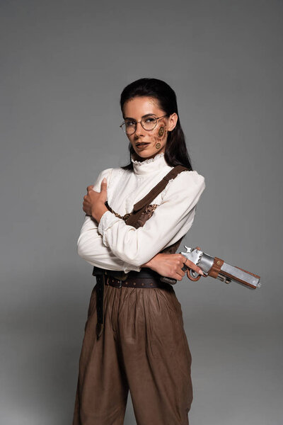 pensive steampunk woman in glasses holding pistol and looking away on grey