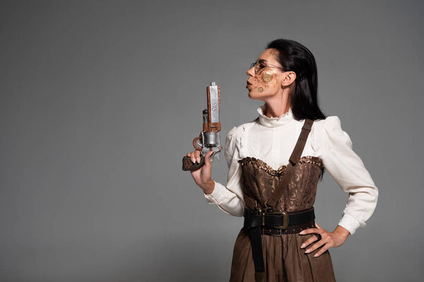 confident steampunk woman standing with hand on hip and holding pistol isolated on grey