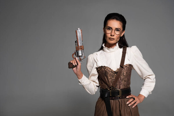 attractive steampunk woman standing with hand on hip and holding pistol isolated on grey