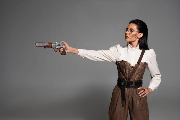 attractive steampunk woman standing with hand on hip and aiming with pistol on grey