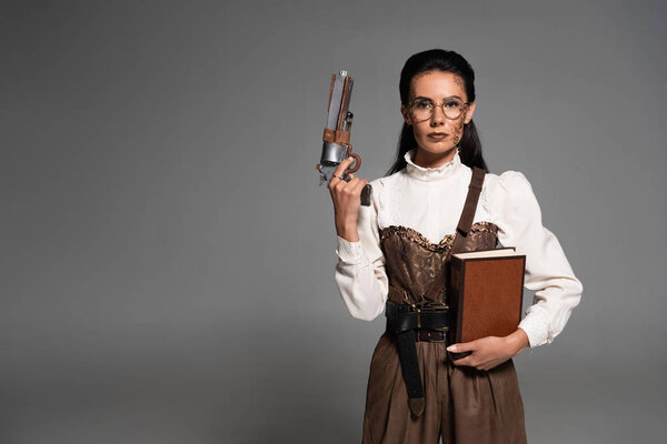 front view of steampunk woman holding book and vintage pistol on grey