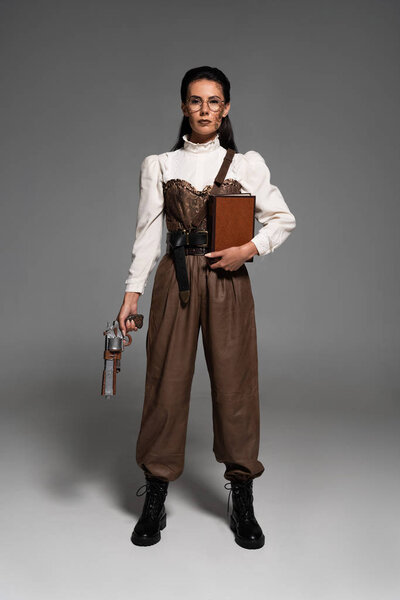 full length view of steampunk woman holding book and vintage pistol on grey