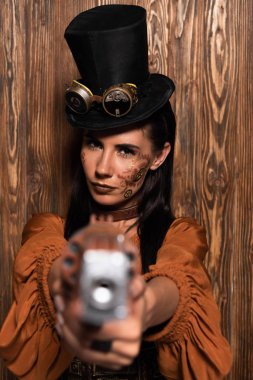 selective focus of serious steampunk woman in top hat with goggles aiming with pistol at camera on wooden clipart