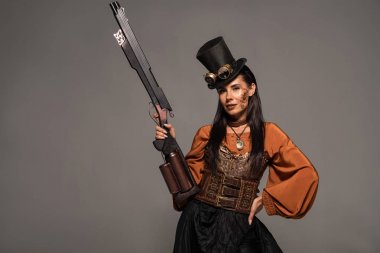 attractive steampunk woman in top hat holding gun and standing with hand on hip isolated on grey clipart