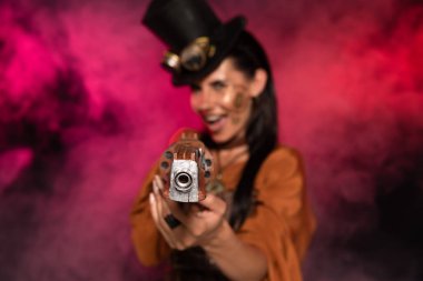 selective focus of screaming steampunk woman aiming with pistol to camera in pink smoke on black clipart