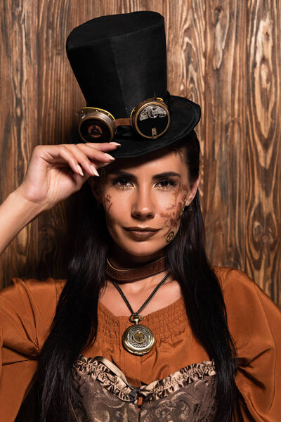 front view of steampunk woman touching top hat with goggles looking at camera on wooden