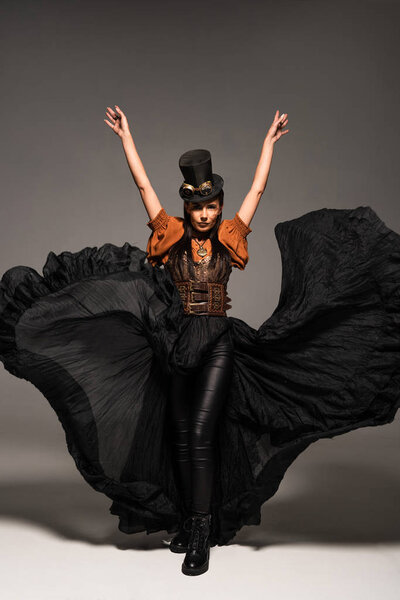 full length view of attractive steampunk woman in top hat with goggles standing with hands up on grey