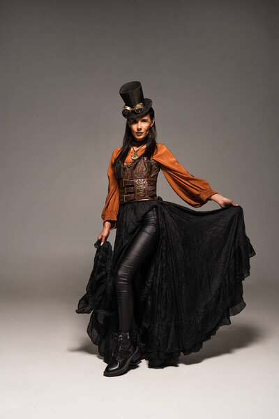 full length view of attractive steampunk woman in top hat with goggles on grey