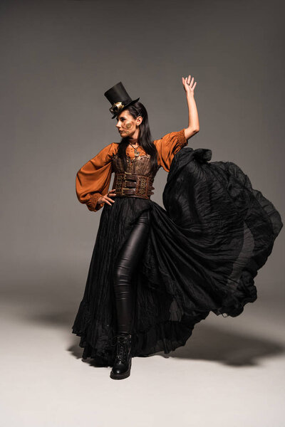 full length view of attractive steampunk woman in top hat with goggles standing with hand on hip on grey
