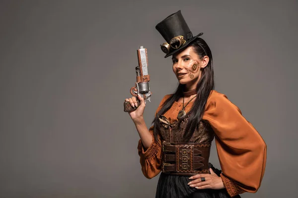 smiling steampunk woman in top hat with goggles standing with hand on hip and holding pistol isolated on grey
