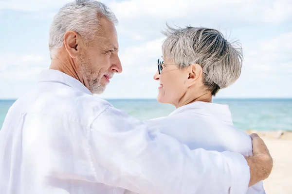smiling senior couple in white shirts embracing and looking at each other near river