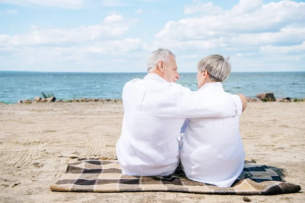back view of senior couple in white shirts sitting on blanket and embracing at beach