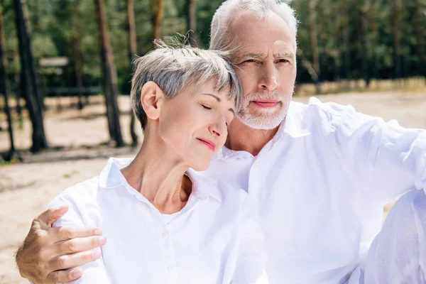 senior couple in white shirts embracing in forest in sunny day