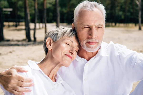 senior couple in white shirts embracing in forest in sunny day