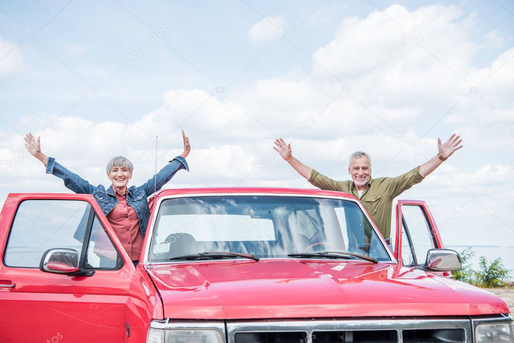 smiling senior couple of tourists standing with hands up near red car at beach