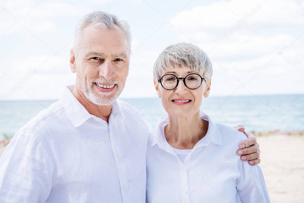 front view of smiling senior couple embracing and looking at camera at beach