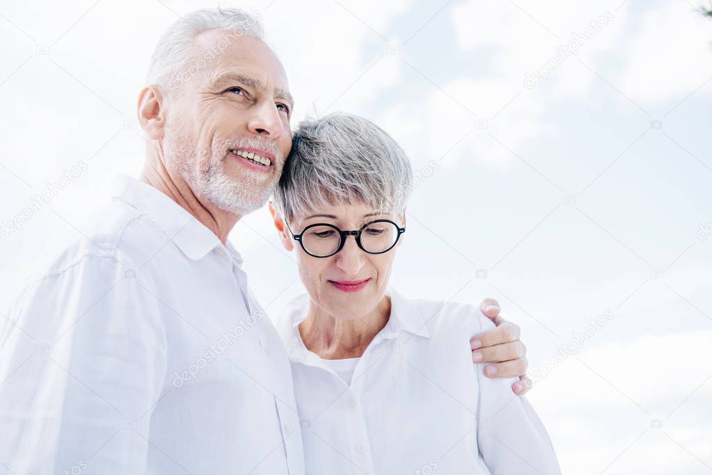 smiling senior couple in white shirts embracing under blue sky