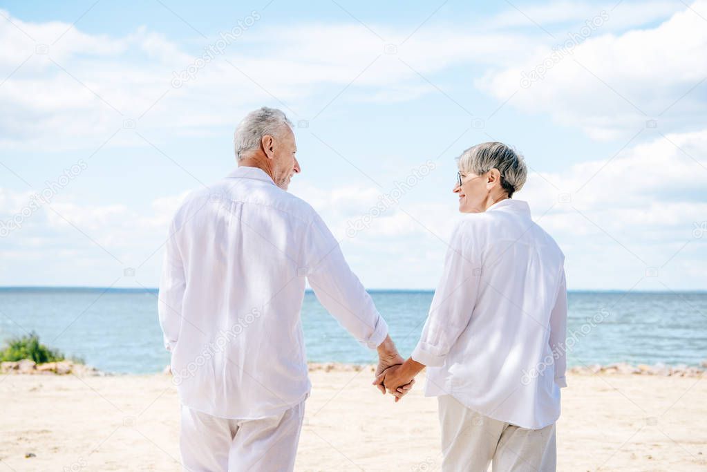 back view of senior couple in white shirts holding hands and looking at each other at beach
