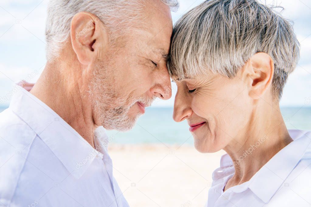 side view of smiling senior couple touching foreheads with closed eyes at beach