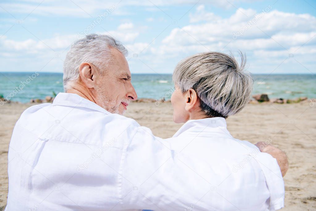 happy senior couple in white shirts embracing and looking at each other at beach
