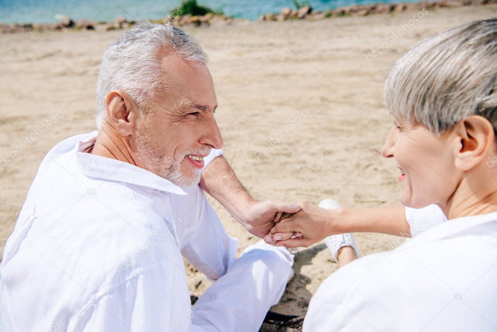 smiling senior couple holding hands and looking at each other at beach