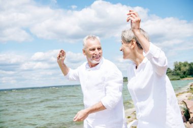 beyaz gömlekli mutlu gülümseyen yaşlı çift birbirlerine bakıyor ve mavi gökyüzü altında plajda gesturing