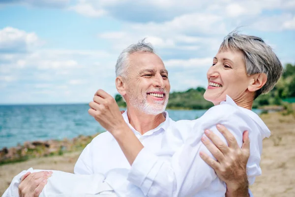 Smiling Senior Man White Shirt Lifting Wife Beach Blue Sky — Stock Photo, Image