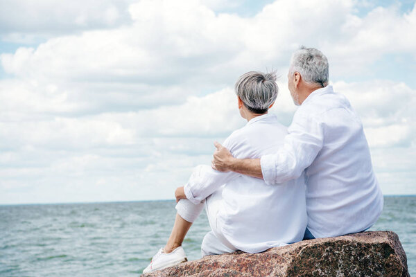back view of senior couple in white shirts sitting on stone and embracing near river