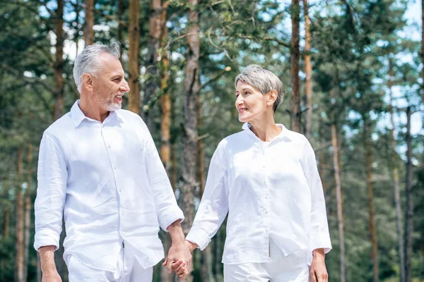 happy senior couple in white shirts holding hands and looking at each other in forest